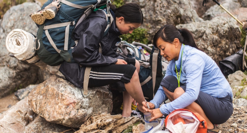 a person wearing backpacking gear sits on a rock while another person helps them on an outward bound course for bipoc